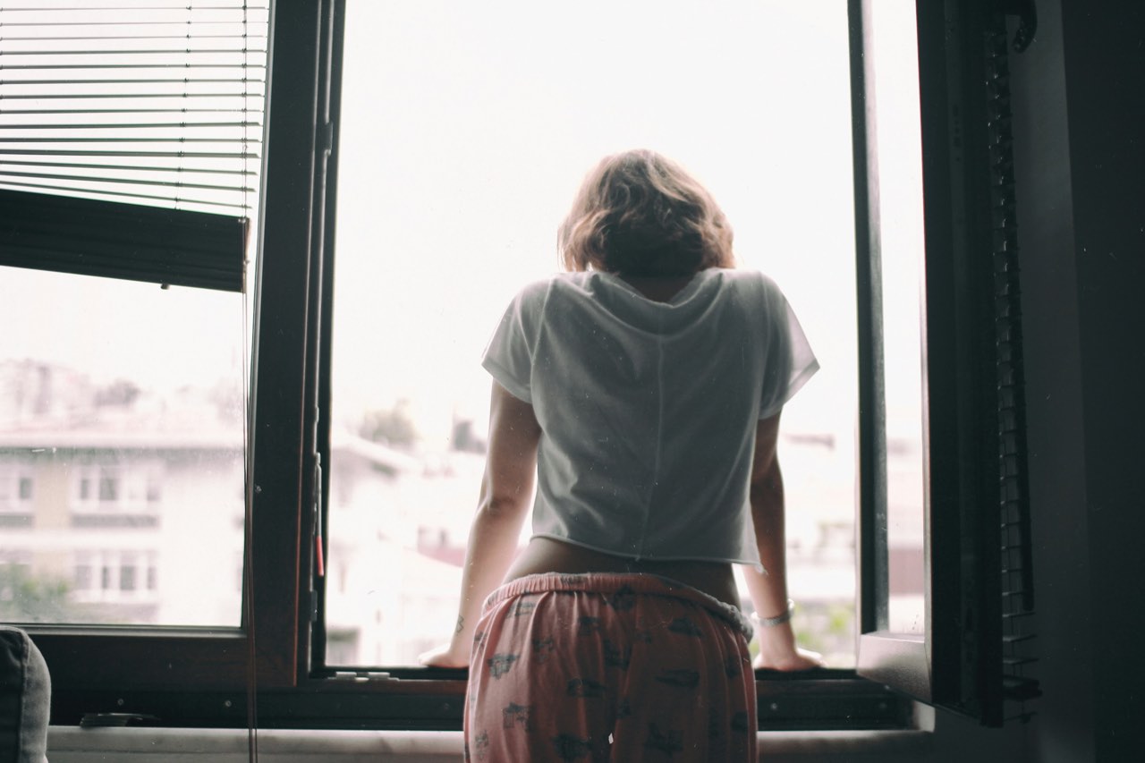 A woman seen from behind, leaning on the sill of an open window in soft daylight.