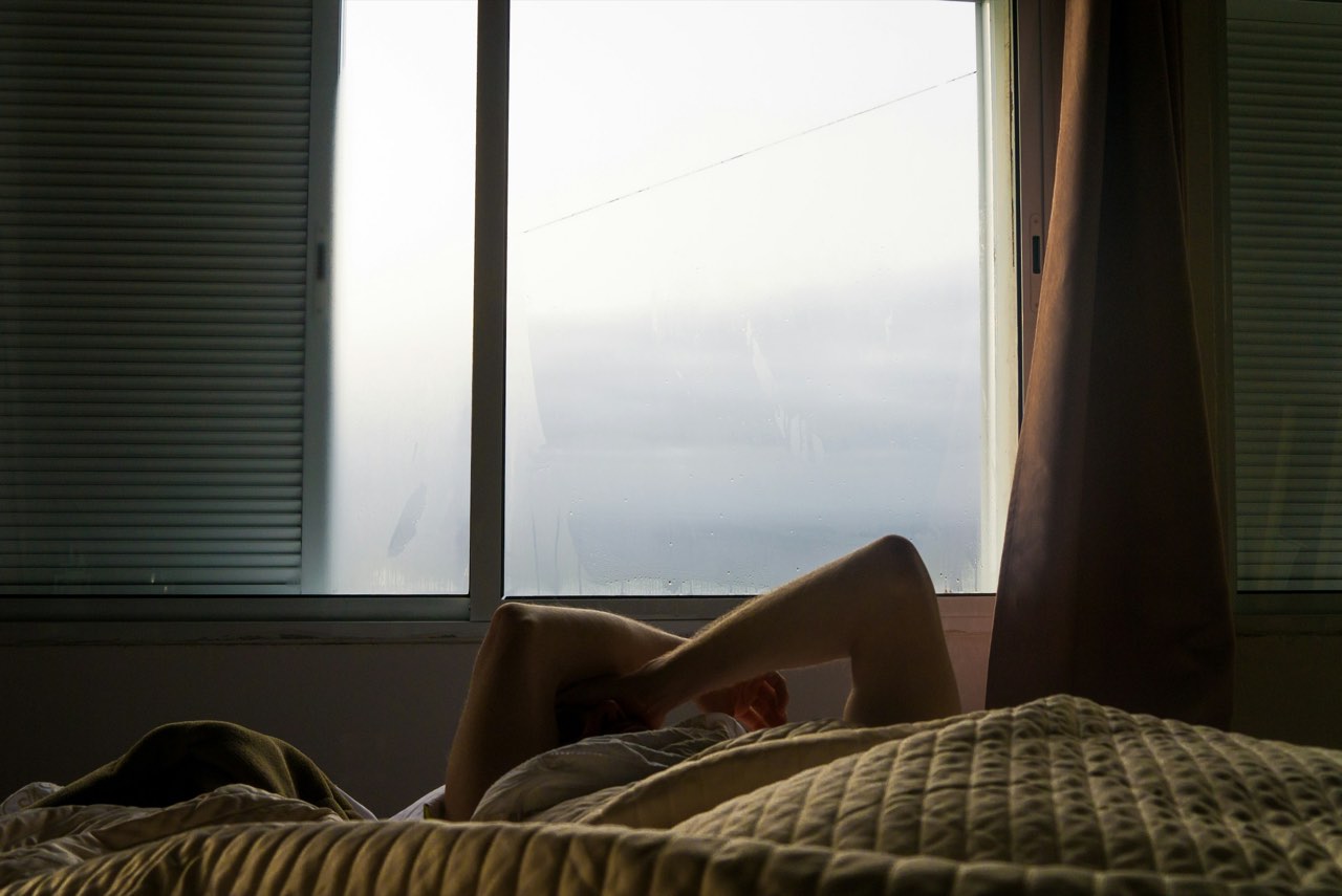 Someone lying in bed at first light, arms raised over their face, beside a misted window.